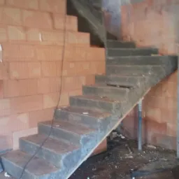 Surowe, betonowe schody wewnętrzne w trakcie budowy domu z czerwonej cegły, widoczny worek gipsu i fragment kolumny z otworem wentylacyjnym.