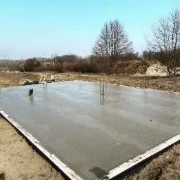 Wylany fundament pod budowę domu z wystającymi prętami zbrojeniowymi, widoczni pracownicy w oddali, teren budowy w słoneczny dzień.