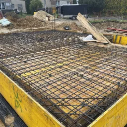 Zbrojenie fundamentów domu jednorodzinnego w deskowaniu, widoczne elementy dystansowe i folia hydroizolacyjna na obrzeżach.