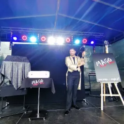 Mężczyzna w złotym fraku prezentuje połączone metalowe obręcze na scenie z oświetleniem, stolikiem z rekwizytami i tablicą reklamową.