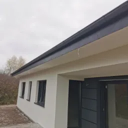 Nowoczesny dom z jasną elewacją, ciemnymi oknami i dachem, widok z dołu na fragment ściany i okna oraz krajobraz w tle.