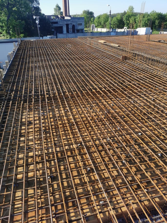 Siatka zbrojeniowa na budowie domu, widok z góry na przygotowaną konstrukcję stropu, w tle powstający budynek z cegły.