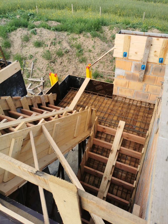 Wykonywanie szalunków pod schody żelbetowe wewnątrz budynku murowanego, widok z góry na zbrojenie i drewniane konstrukcje, w tle zielona łąka.