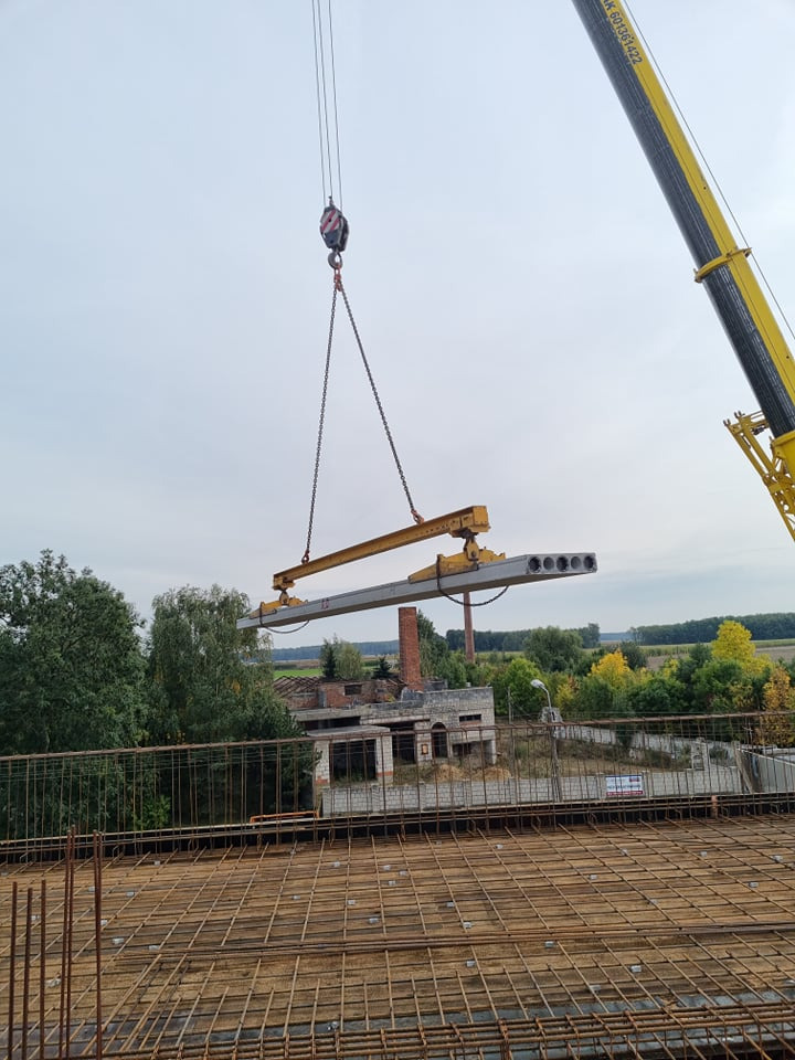 Żuraw transportuje prefabrykowany strop kanałowy nad zbrojeniem, w tle zarośnięty budynek i zielona roślinność.