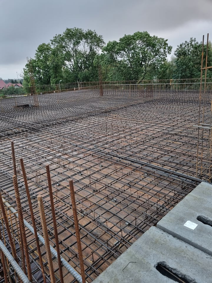 Zbrojenie stropu domu jednorodzinnego z widocznymi elementami szalunku i wystającymi prętami, na tle zieleni drzew i pochmurnego nieba.