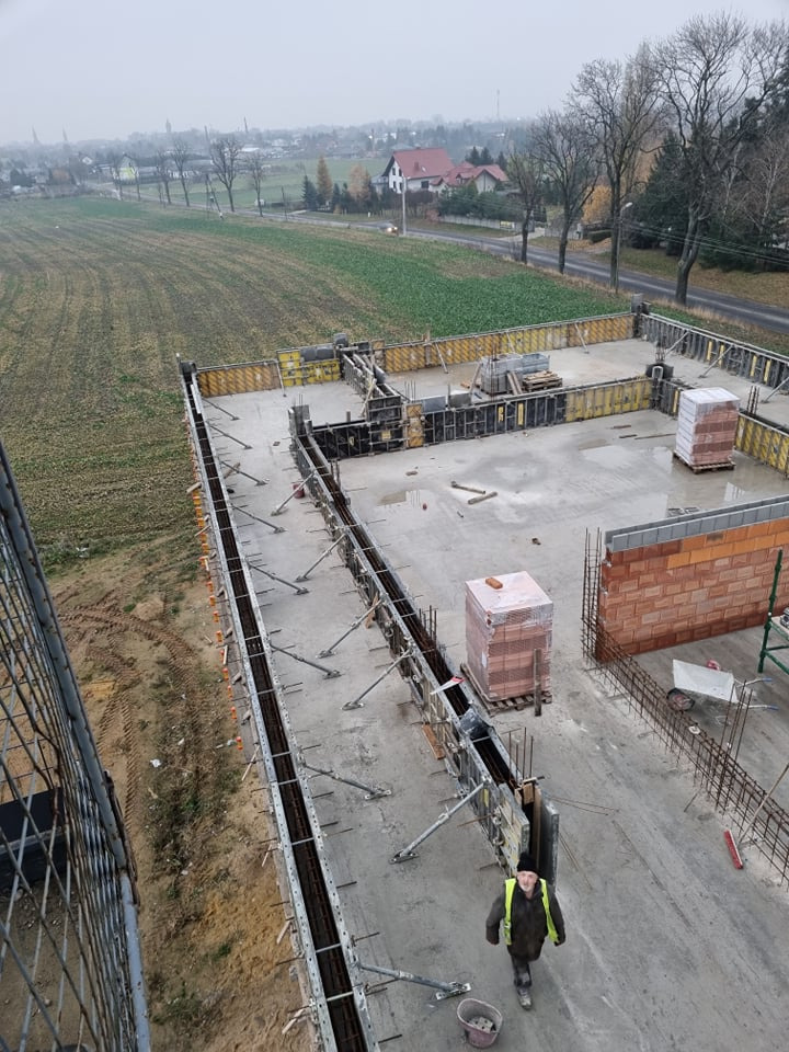 Fundamenty domu jednorodzinnego w trakcie budowy, widok z góry na szalunki, zbrojenie i częściowo wymurowane ściany, na tle jesiennego krajobrazu.