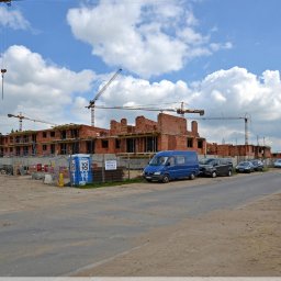 Optima Business - Budowa bloku z czerwonej cegły w słoneczny dzień. Widoczne rusztowania, żurawie i samochody. Okna w trakcie montażu.