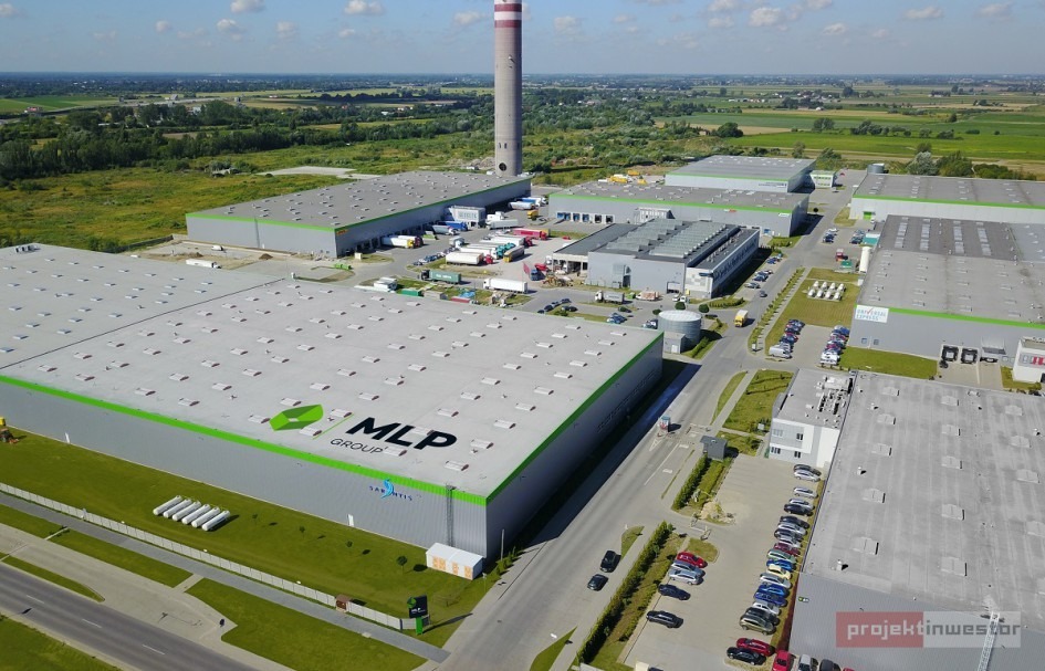 Widok z lotu ptaka na kompleks hal magazynowych z szarymi dachami i zielonymi akcentami, z parkingami i drogami dojazdowymi. W tle komin i zielone tereny.