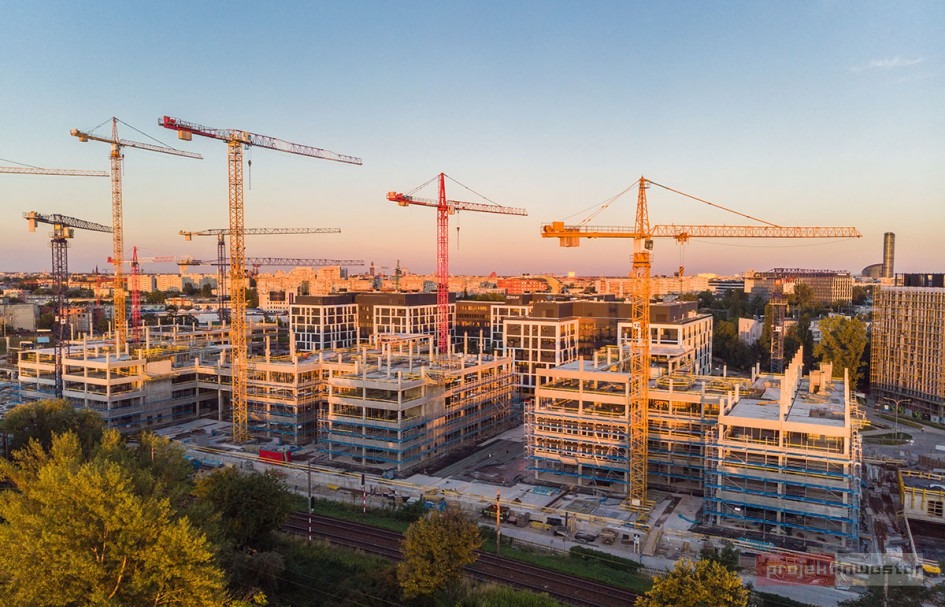 Widok z lotu ptaka na budowę osiedla mieszkaniowego z licznymi żurawiami na tle zachodzącego słońca i panoramy miasta.
