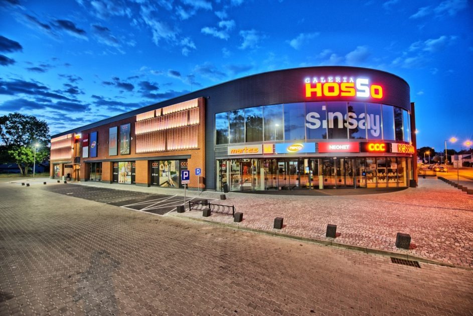 Oświetlona fasada Galerii Hosso o zmierzchu, z dużymi witrynami sklepów Sinsay, Martes i CCC. Widoczny parking z kostki brukowej i znak P.