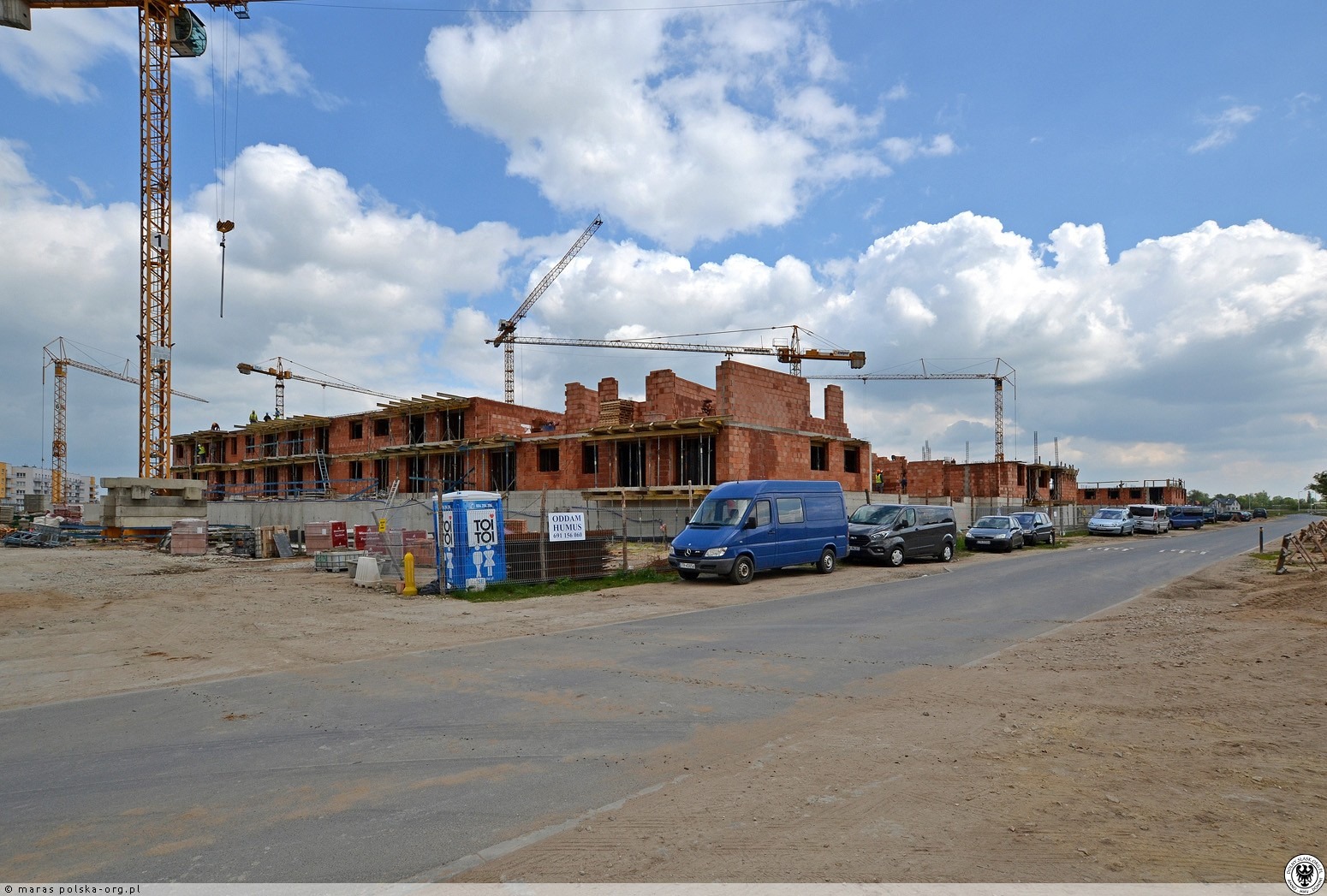 Budowa bloku z czerwonej cegły w słoneczny dzień. Widoczne rusztowania, żurawie i samochody. Okna w trakcie montażu.