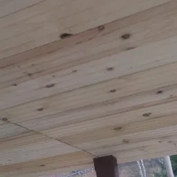 Drewniany sufit z widocznymi belkami i deskami, z perspektywy z dołu, widoczne fragmenty otoczenia zewnętrznego.