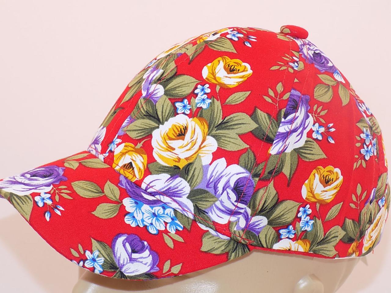 Czerwona czapka z daszkiem w kwiaty, z motywem róż w kolorach żółtym, białym i fioletowym, umieszczona na manekinie.
