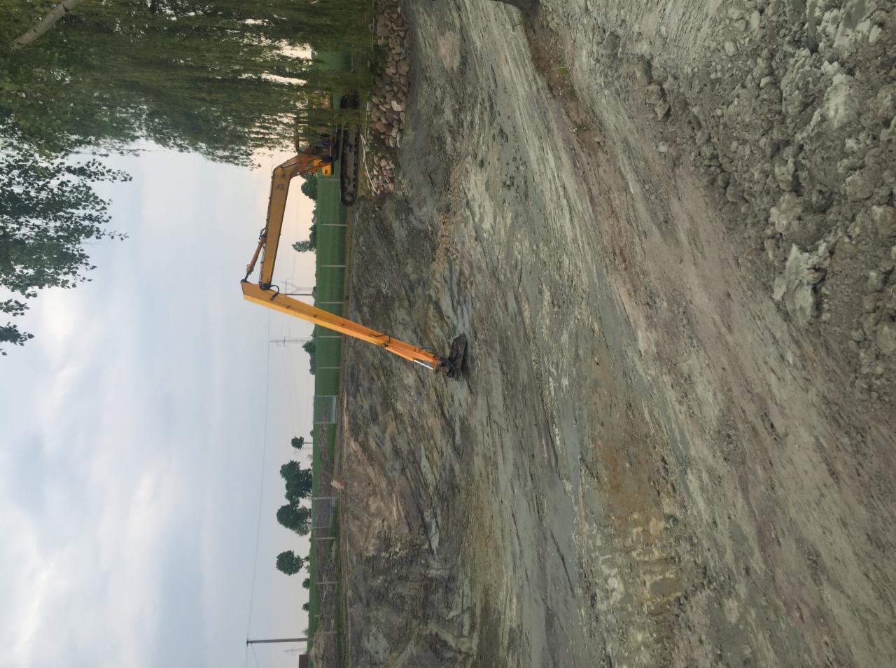 Żółta koparka na placu budowy, widoczne świeżo wykopane rowy w ziemi, w tle drzewa i słupy energetyczne.