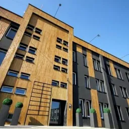 Nowoczesny budynek biurowy z fasadą w odcieniach szarości i drewna, z licznymi oknami i dekoracyjnymi kulami roślinnymi w donicach przed wejściem.