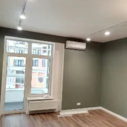 Wnętrze pokoju po odświeżeniu: ściany pomalowane na oliwkowy kolor, drewnopodobne panele na podłodze, białe listwy przypodłogowe, grzejnik pod oknem balkonowym, klimatyzator na ścianie...