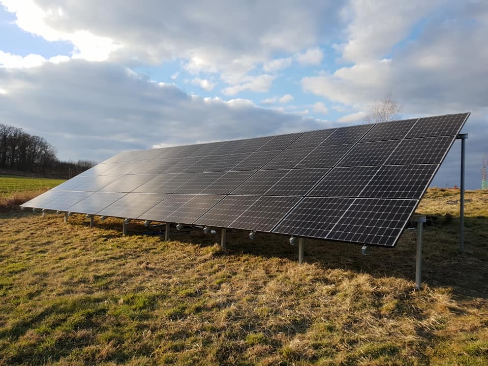 Instalacja paneli słonecznych na metalowej konstrukcji, widok z boku na łące z drzewami w tle, pochmurne niebo.
