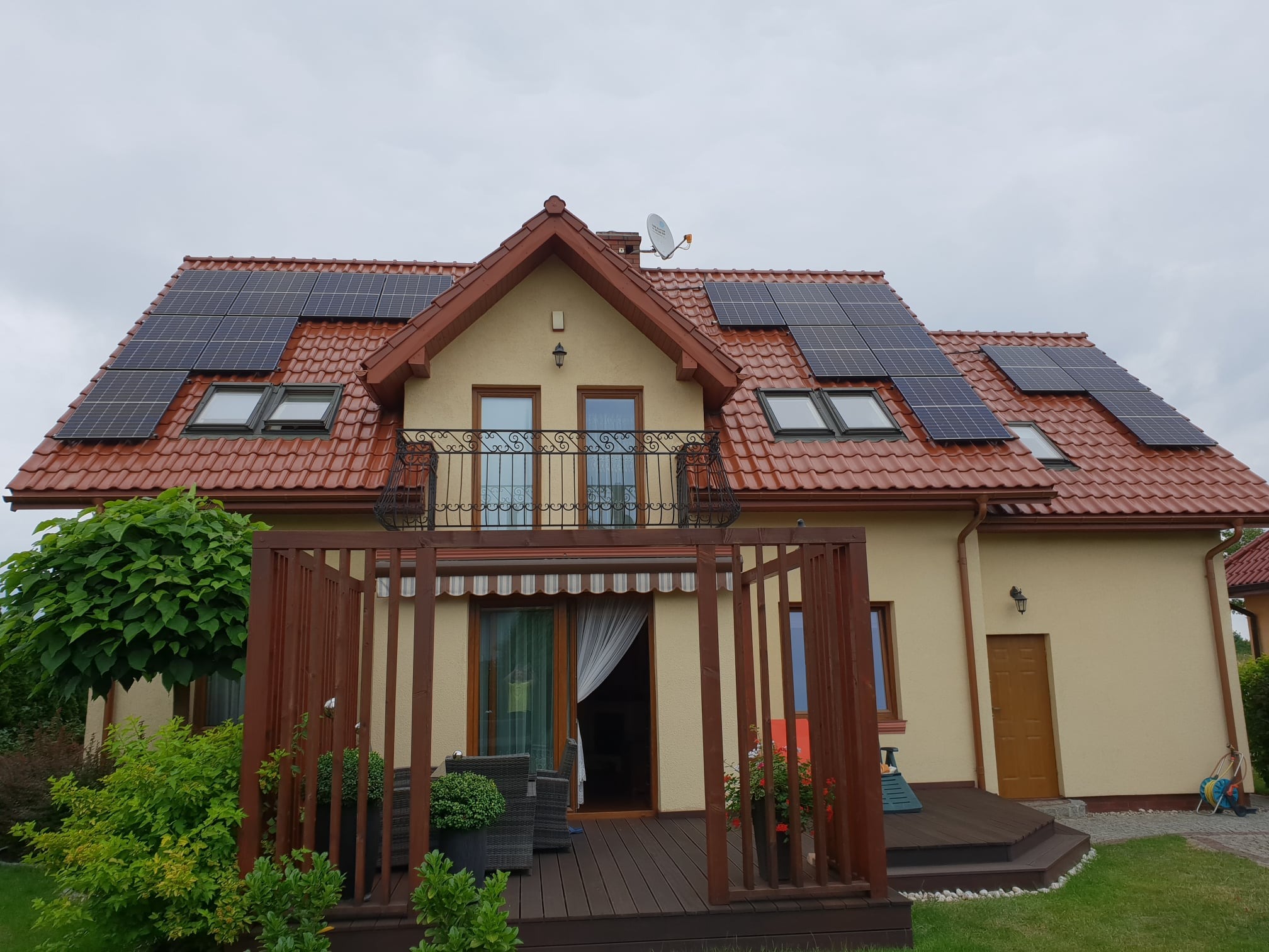 Dom jednorodzinny z panelami słonecznymi na dachu z czerwonej dachówki, widoczne okna dachowe, balkon z kutą balustradą i drewniany taras.