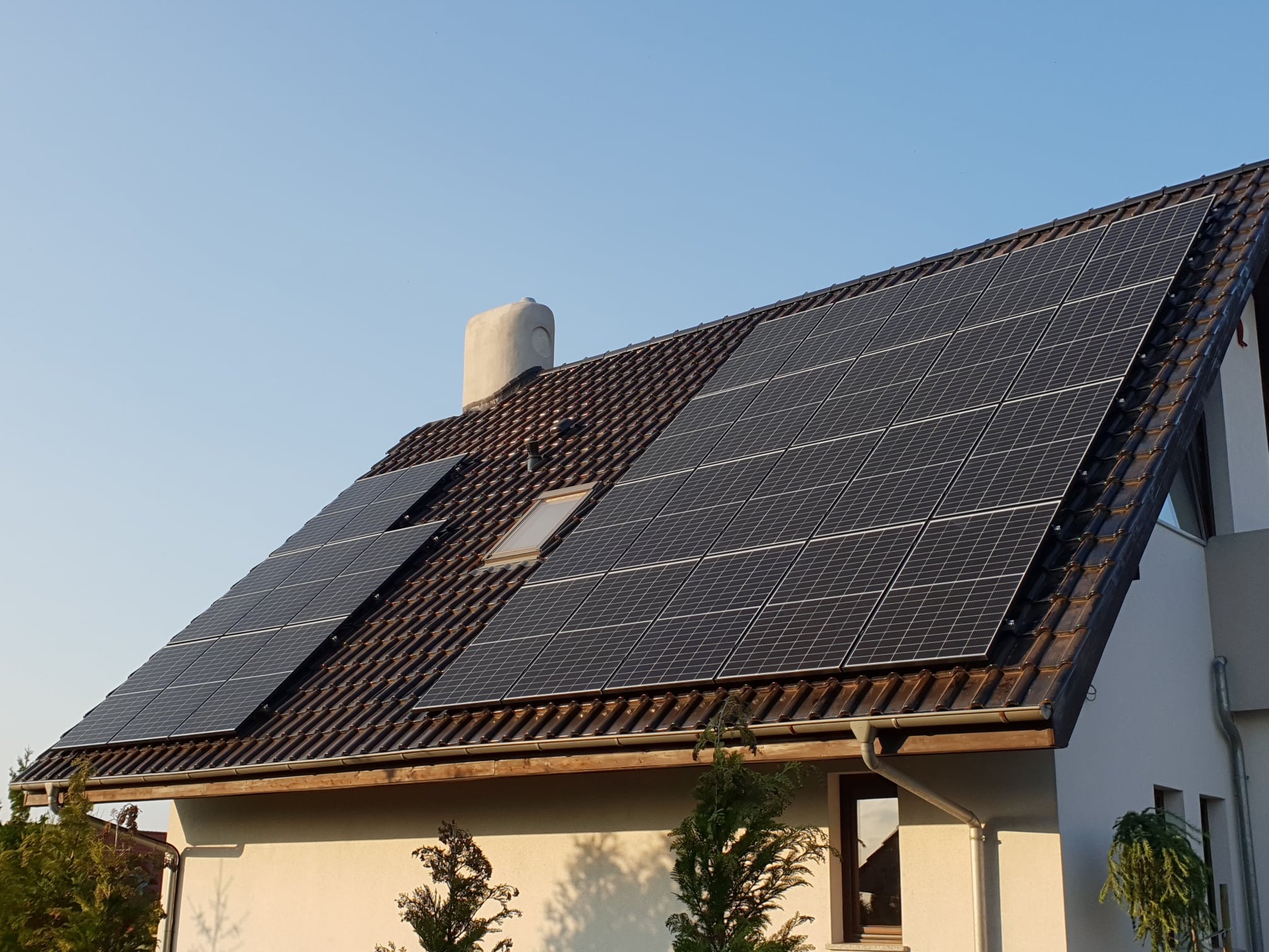 Panele słoneczne na brązowym dachu domu jednorodzinnego z kominem i oknem dachowym, widok z dołu w słoneczny dzień.