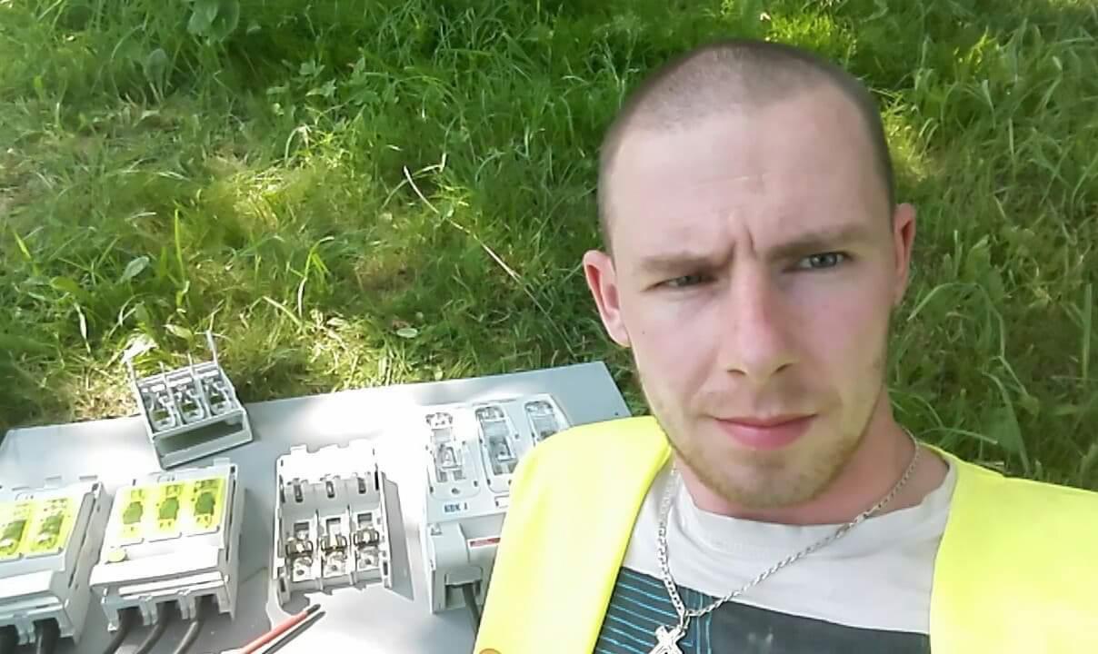 Mężczyzna w kamizelce odblaskowej pozuje na tle rozłożonej skrzynki z bezpiecznikami i zielonej trawy. Widoczne elementy instalacji elektrycznej.