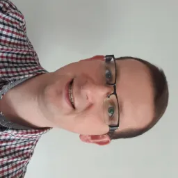 Uśmiechnięty mężczyzna w okularach i koszuli w kratę, patrzy w obiektyw, jasne tło, zdjęcie obrócone o 90 stopni w lewo.