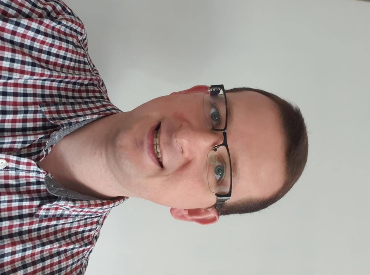 Uśmiechnięty mężczyzna w okularach i koszuli w kratę, patrzy w obiektyw, jasne tło, zdjęcie obrócone o 90 stopni w lewo.