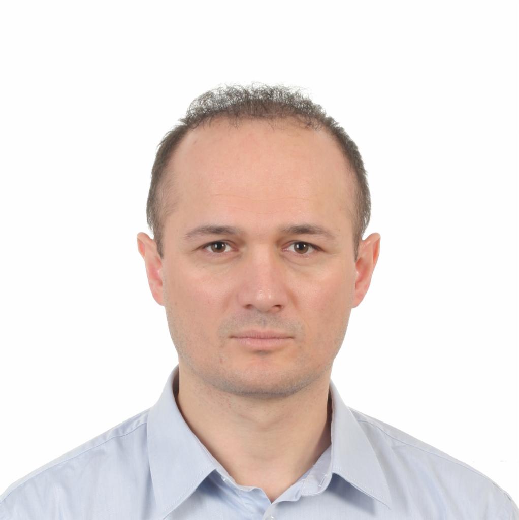 Portret mężczyzny w jasnoniebieskiej koszuli na białym tle, z wyraźnymi zmarszczkami na czole i krótkimi, ciemnymi włosami.