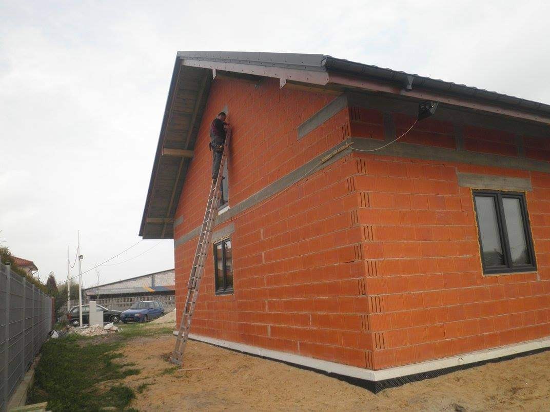 Prace na elewacji domu z czerwonej cegły ceramicznej, pracownik na drabinie, widoczne okna i fragment dachu z orynnowaniem, surowy stan wykończenia.