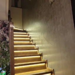 Drewniane schody z podświetleniem LED na każdym stopniu, oświetlające drogę w górę, obok ściana z dekoracyjnym tynkiem i kinkietem oraz ściana z tapetą w kwiaty.