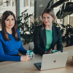 Dwie prawniczki siedzą przy biurku z laptopem i kubkami, zielone rośliny w tle, kobieta w niebieskiej bluzce trzyma kubek.