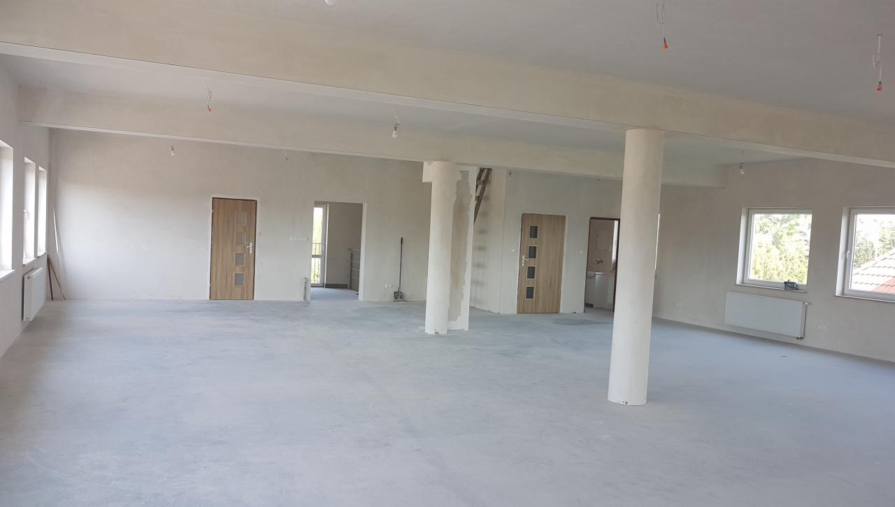 Surowe wnętrze biurowe przed wykończeniem, widoczne kolumny, ściany przygotowane pod gładzie, surowy beton na podłodze, instalacja elektryczna częściowo wyprowadzona, okna z widokiem na zieleń.