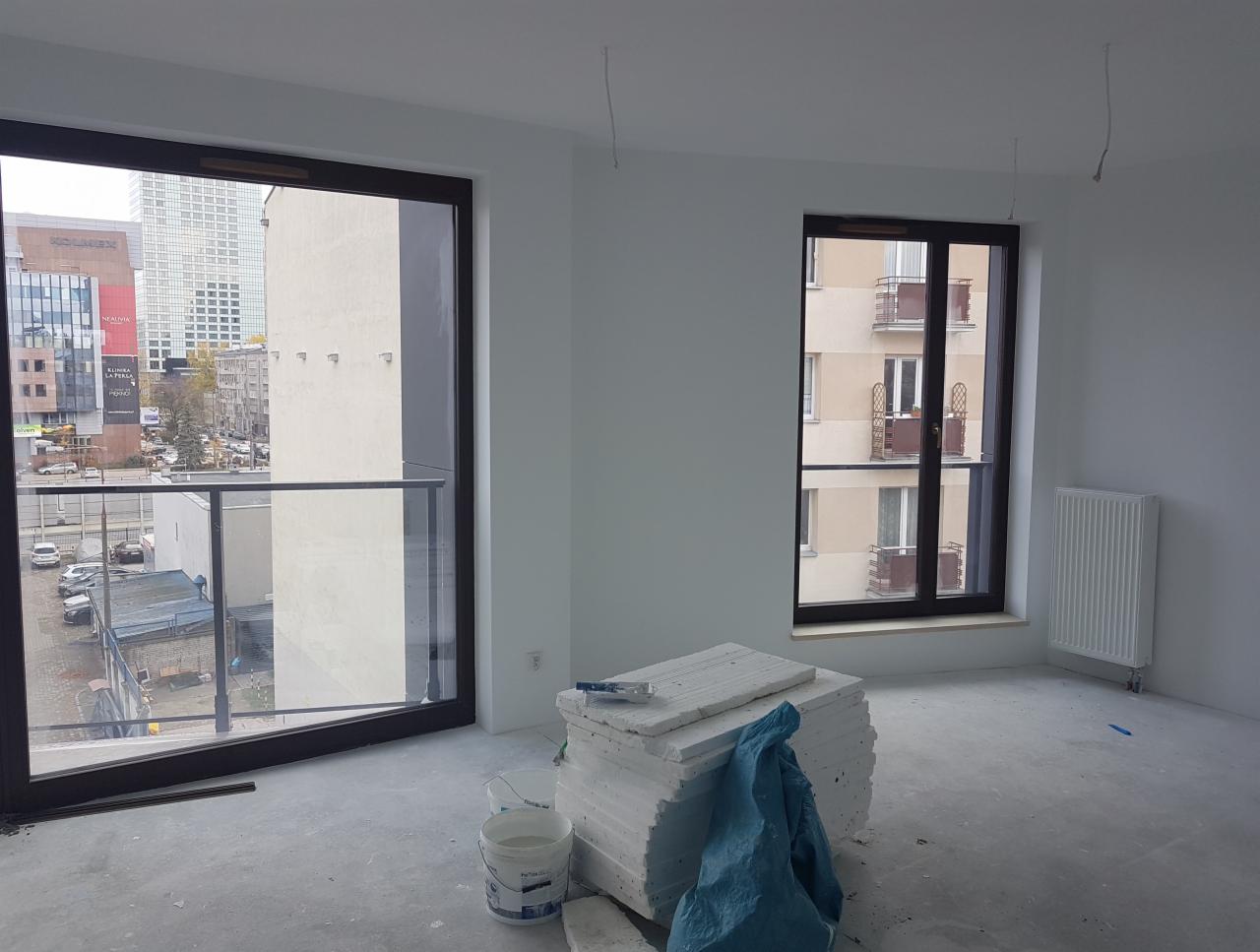 Wnętrze pokoju po świeżym pomalowaniu na biało, widoczne dwa okna z widokiem na miasto oraz blok mieszkalny, na podłodze leżą płyty styropianowe przykryte niebieską folią i wiadro z farbą.