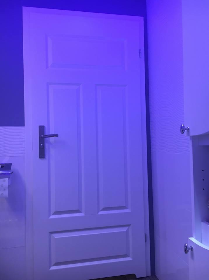 Białe, panelowe drzwi wewnętrzne z klamką w łazience oświetlonej fioletowym światłem. Widoczny fragment ściany z dekoracyjną teksturą oraz narożnik szafki łazienkowej.
