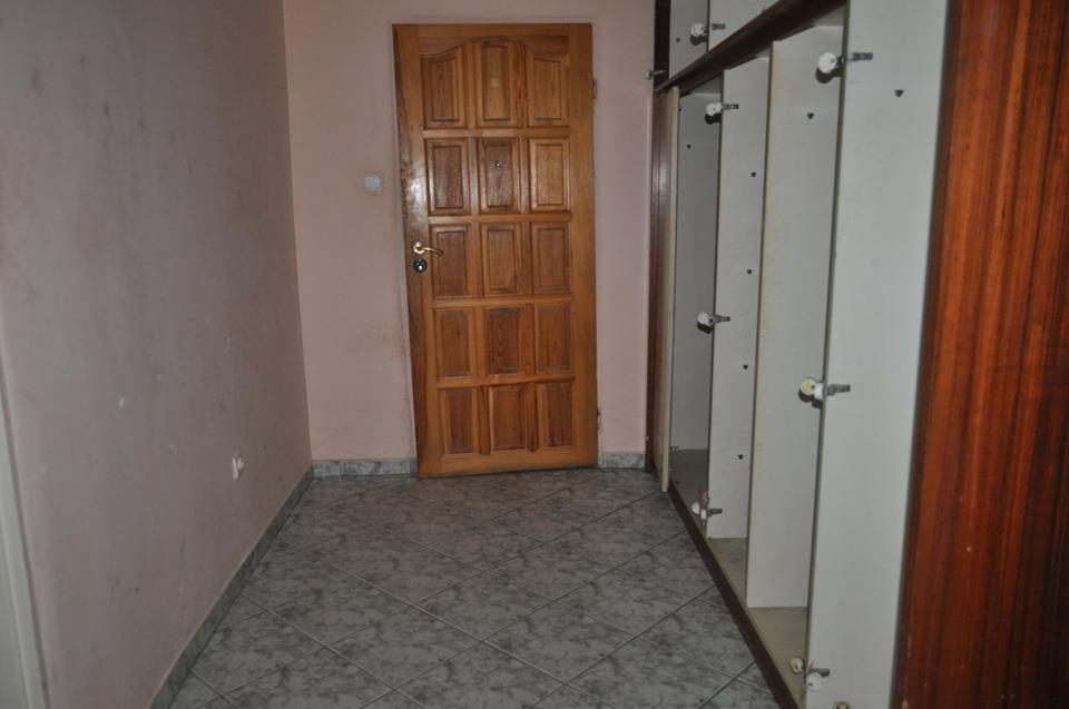 Wnętrze korytarza z drewnianą okładziną drzwiową i częściowo zmontowaną szafą wnękową z białymi półkami, widoczne szare płytki podłogowe.