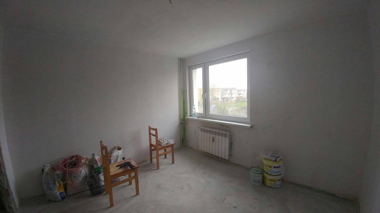 Pokój w trakcie remontu: surowe ściany po gładzi, niepoziomowana podłoga, grzejnik pod oknem, worki z materiałami budowlanymi i dwa krzesła w narożniku.