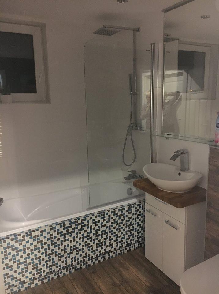 Wykończona łazienka z wanną obudowaną mozaiką w odcieniach bieli, niebieskiego i brązu, kabiną prysznicową ze szklanym panelem i umywalką nablatową na szafce z drewnianym blatem.