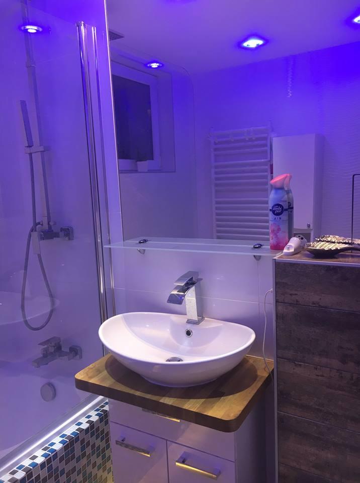 Nowoczesna łazienka z owalną umywalką na drewnianym blacie, chromowaną baterią i kabiną prysznicową z mozaiką, oświetlona fioletowym światłem LED.