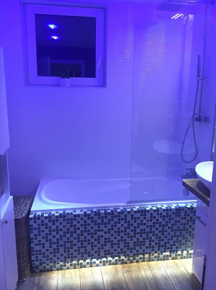 Nowoczesna łazienka w niebieskiej tonacji: wanna obudowana mozaiką, drewniana podłoga, szklana kabina prysznicowa i oświetlenie LED.