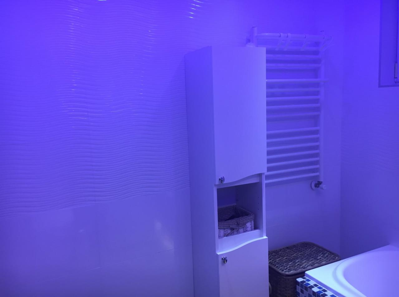 Nowoczesna łazienka z białymi meblami podświetlona niebieskim światłem, widoczna szafka łazienkowa z koszem wiklinowym, grzejnik drabinkowy oraz fragment wanny z mozaiką.