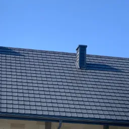 Szary dach z blachodachówki z dwoma kominami na tle błękitnego nieba, widoczne orynnowanie i fragment ściany budynku w stanie surowym.