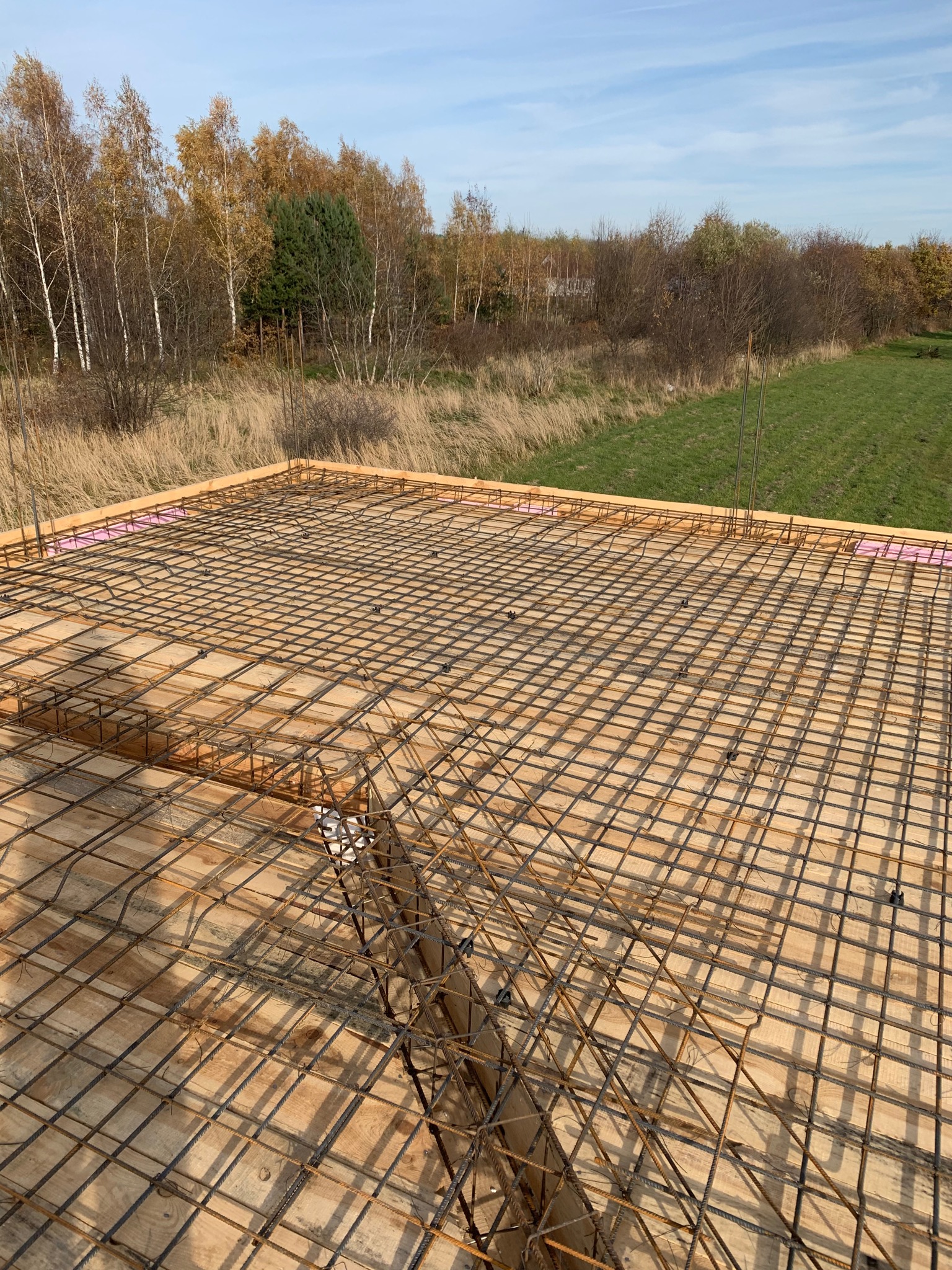 Zbrojenie płyty fundamentowej na drewnianym szalunku, widok z góry na siatkę stalową, różowy styropian na obrzeżach, w tle jesienny krajobraz z drzewami i polami.