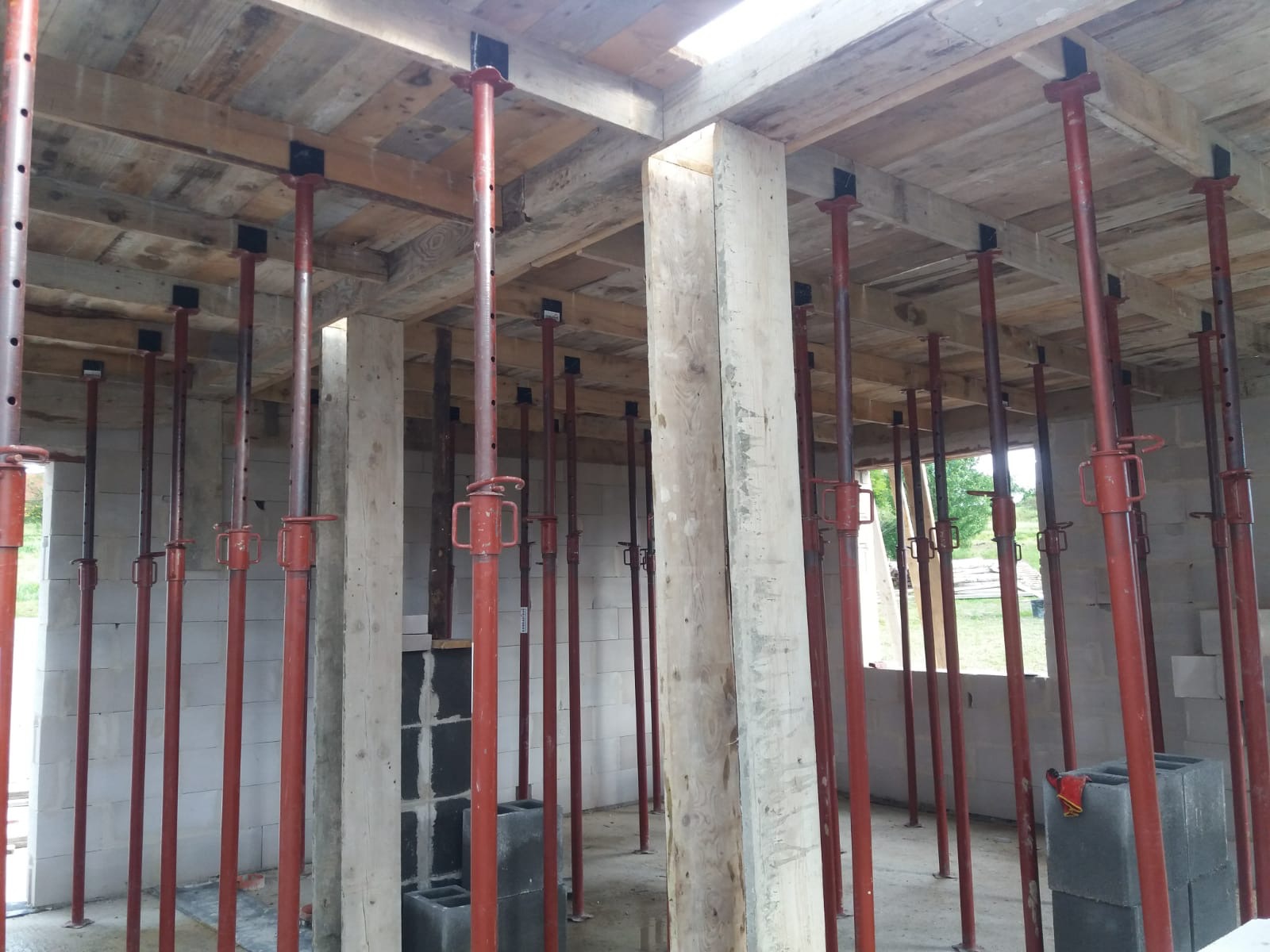 Wnętrze budynku w trakcie budowy z drewnianym stropem podpartym czerwonymi stemplami budowlanymi i ścianami z bloczków betonowych.