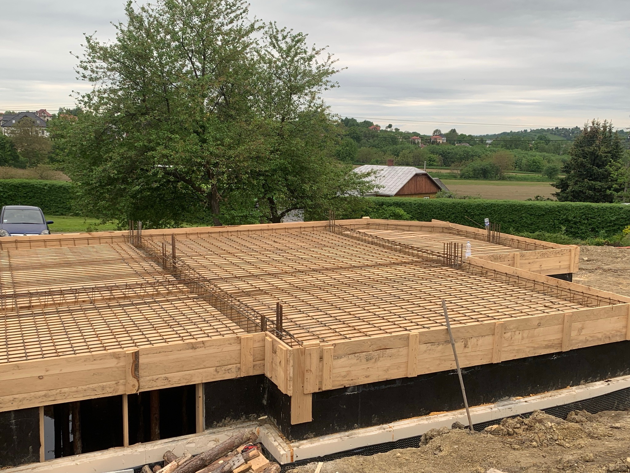 Zbrojenie płyty fundamentowej z drewnianym szalunkiem na tle zieleni i pochmurnego nieba.
