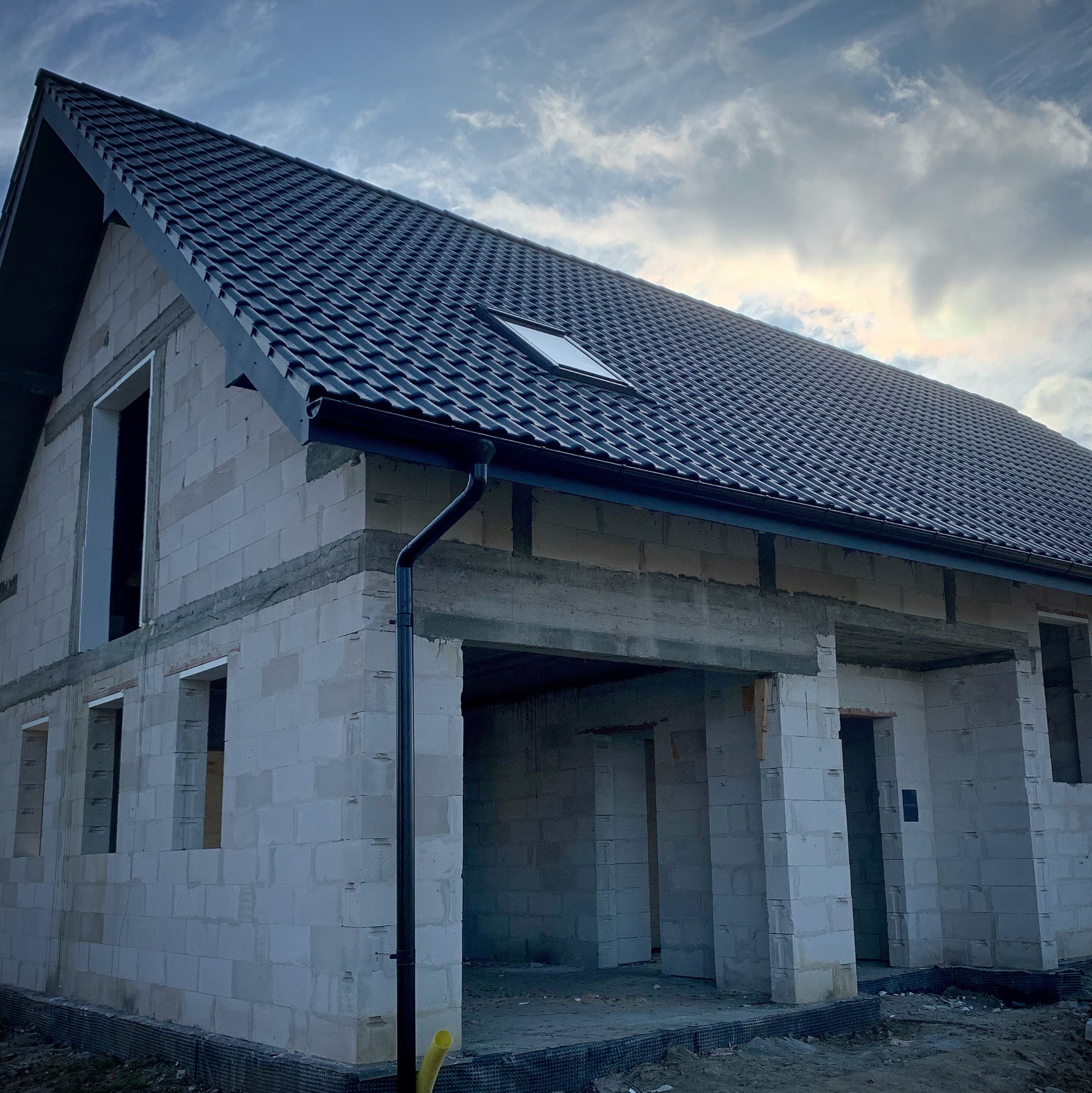 Surowy stan domu w trakcie budowy z białych bloczków, z ciemną dachówką i czarną rynną, widoczne niezadaszone otwory okienne i drzwiowe, na tle pochmurnego nieba.