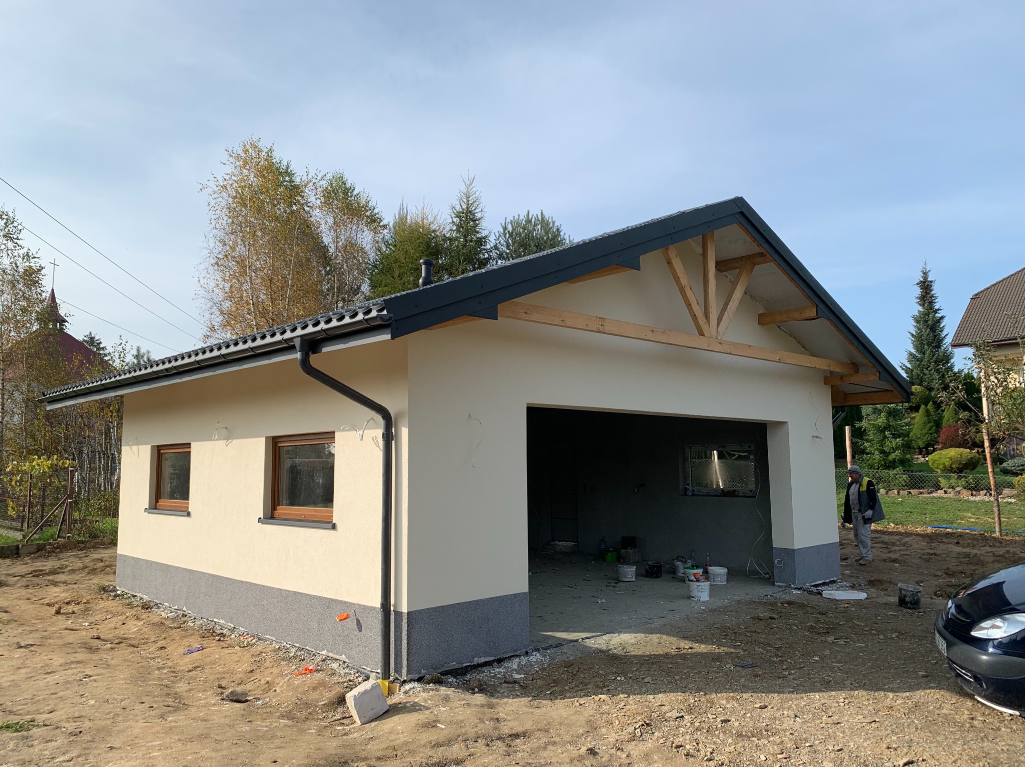 Budynek garażowy w trakcie budowy, z widoczną surową konstrukcją dachu, otynkowaną elewacją w odcieniach beżu i szarości, otoczony ziemią i częściowo zaparkowanym samochodem.