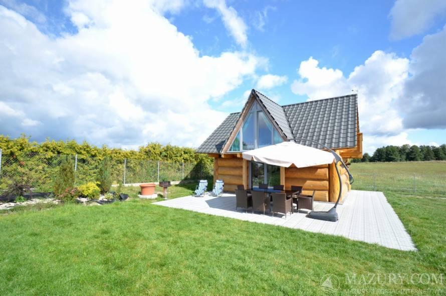 Drewniany domek letniskowy z dużym oknem, tarasem z meblami ogrodowymi i parasolem, otoczony zielonym trawnikiem i ogrodzeniem z tujami, na tle błękitnego nieba z chmurami.