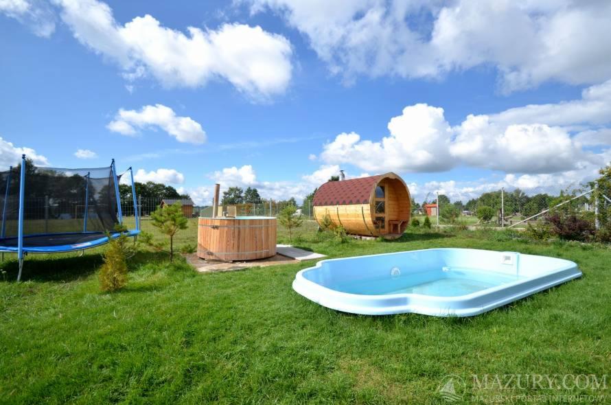 Zewnętrzne spa z drewnianą sauną w kształcie beczki, balią i basenem na zielonym trawniku pod błękitnym niebem z chmurami, widoczny także trampolina.