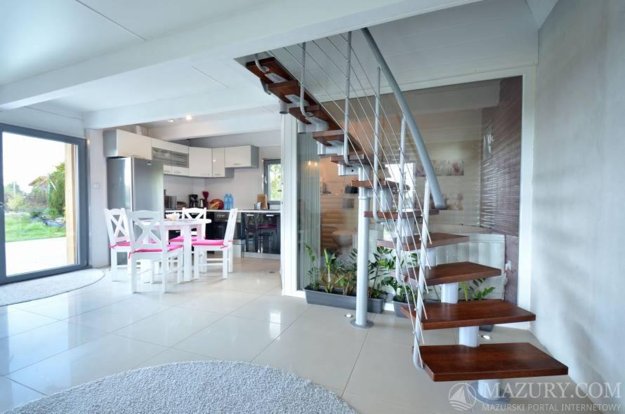 Jasne wnętrze z nowoczesnymi schodami spiralnymi z drewnianymi stopniami, widok na jadalnię z białym stołem i krzesłami z różowymi poduszkami, oraz kuchnię w tle.