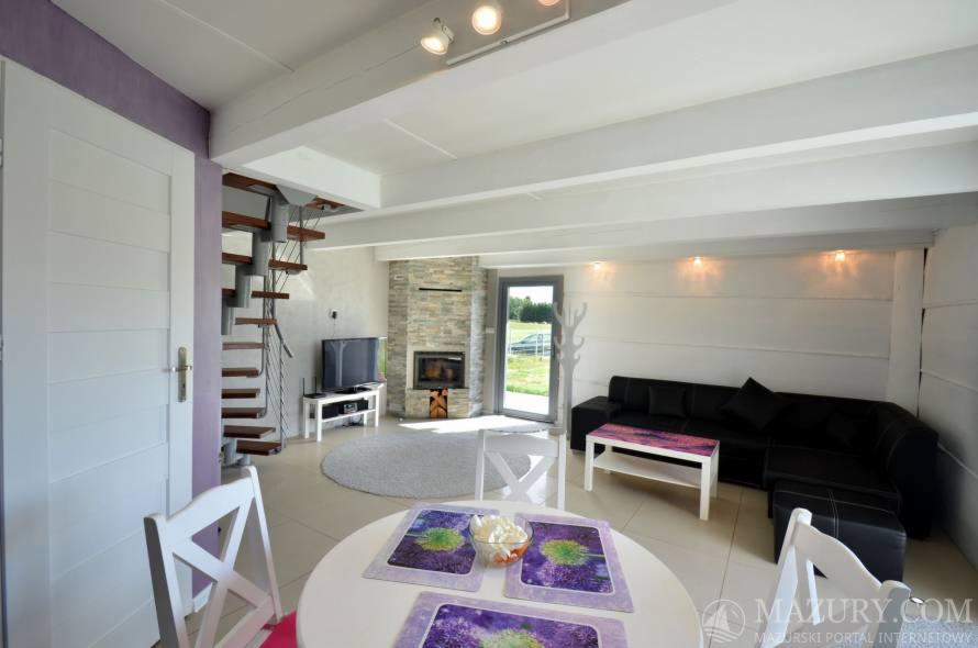 Jasne wnętrze salonu z białymi ścianami, spiralnymi schodami, kominkiem obłożonym kamieniem, czarnym narożnikiem i okrągłym stołem z dekoracyjnymi podkładkami. Widoczne drzwi balkonowe z widokiem...