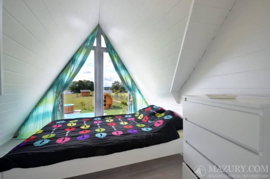 Sypialnia na poddaszu z oknem w kształcie trójkąta, widok na ogród z jacuzzi i sauną, pościel w kolorowe usta, białe ściany z boazerią, komoda w rogu.
